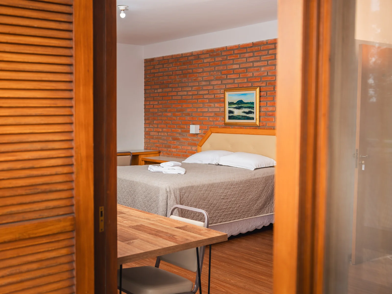 Cama de casal da Suíte Família vista pela porta da sacada, com parede de tijolos e quadro